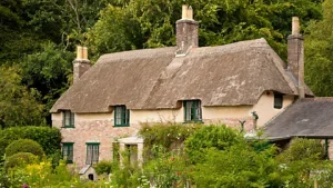 Historic brick thatched cottage with green window frames surrounded by lush English greenery.